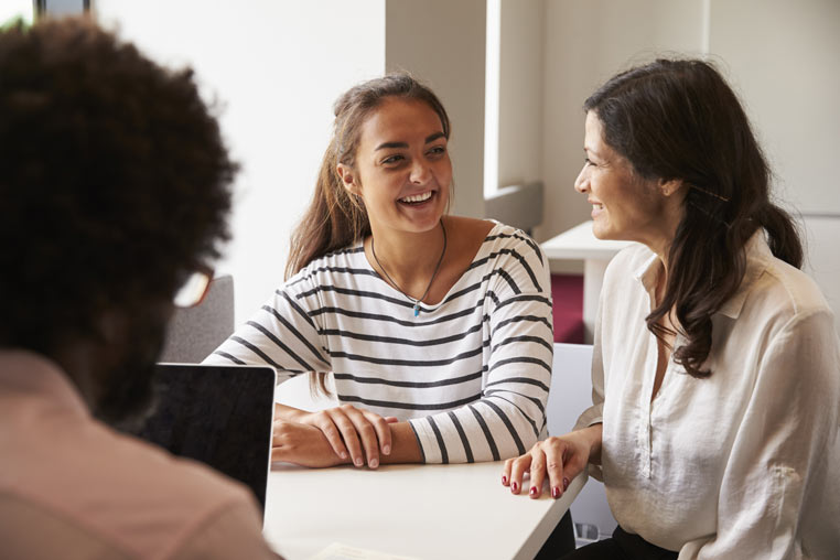 A teenage girl smiles in conversation with an adult seated at a table..